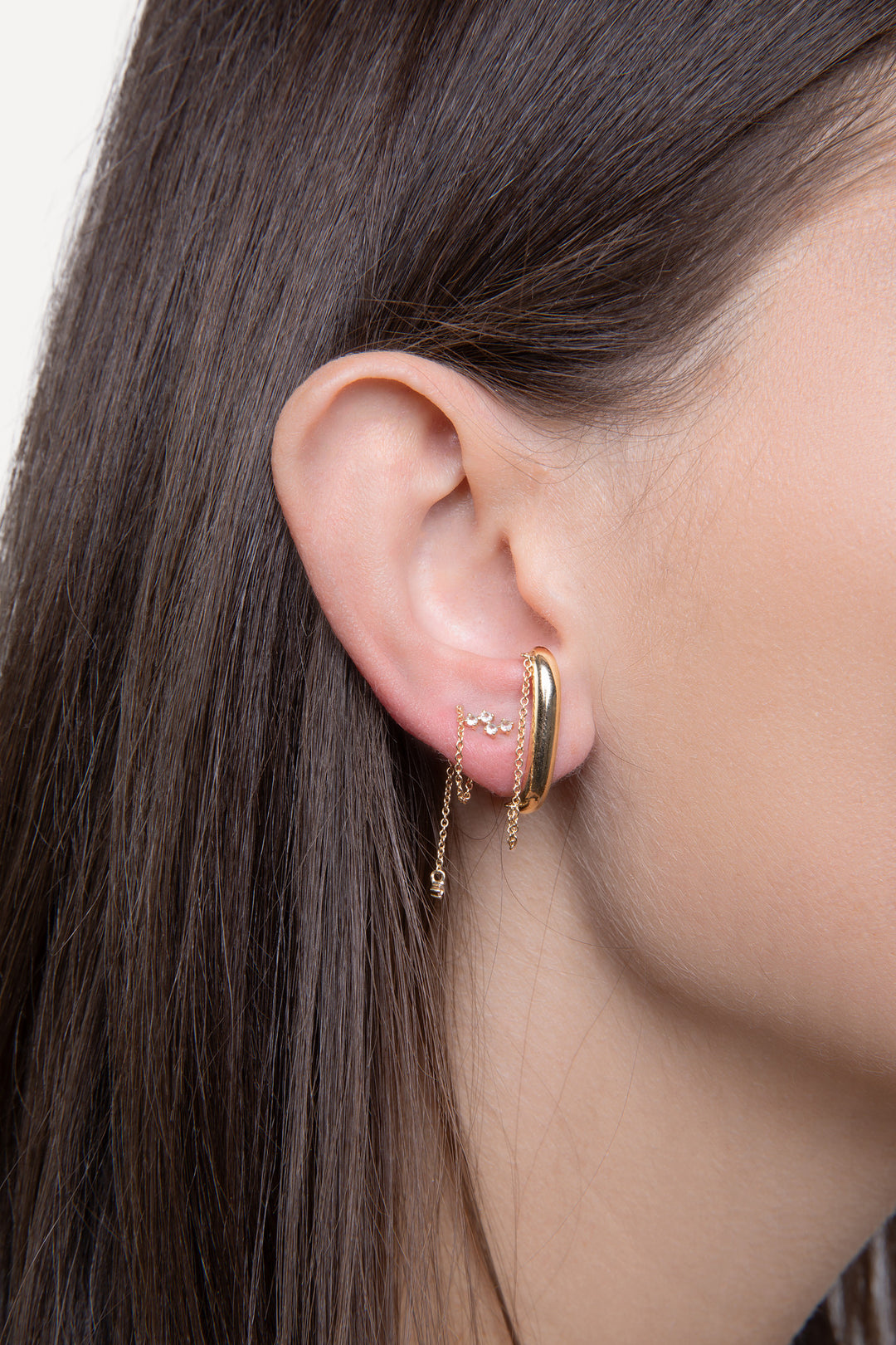 n image of a woman’s ear with two earrings. The first earring is a solid gold ear suspender with chain details and the second is a dangling chain earring with diamond accents