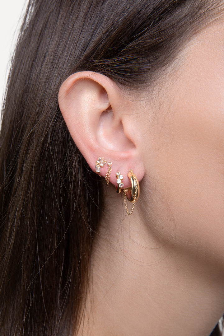 n image of a woman’s ear with four earrings. The first earring is a solid gold hoop earring with chain detailing then a smaller hoop with diamonds and then chain and diamonds studs.