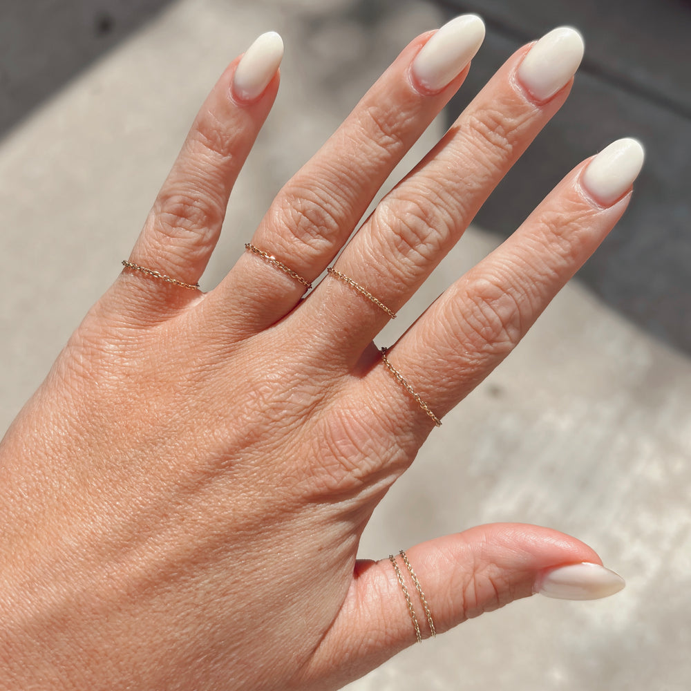 Hand with beige nail polish wearing skinny stacking chain gold rings on a neutral background