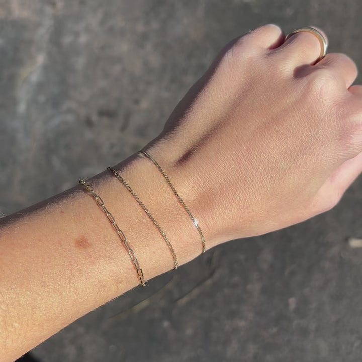 video of a womans wrist wearing three solid gold bracelets. Paperclip, Valentino, Figaro & Single Curb styles.